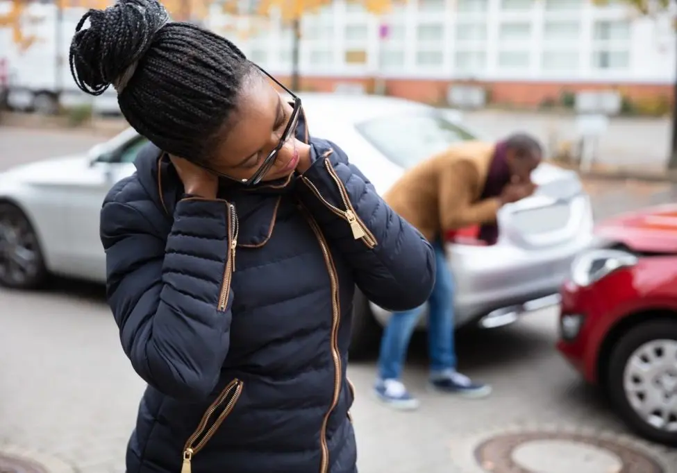A young woman holds the back of her neck after a fender-bender while the other driver looks at the damage