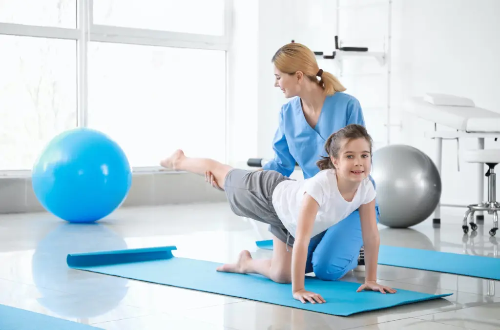 A smiling therapist guides a young girl through a balance exercise on a blue mat in a bright room